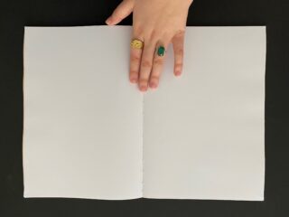 Eine Hand mit zwei Ringen ruht auf einem offenen, leeren, weißen Notizbuch vor schwarzem Hintergrund.