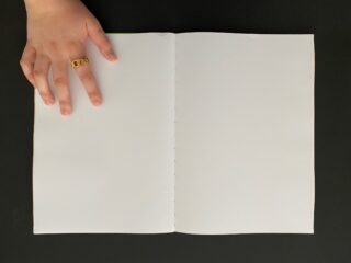 Eine Hand mit einem goldenen Ring ruht auf der oberen linken Ecke eines offenen, leeren Buches vor einem schwarzen Hintergrund.