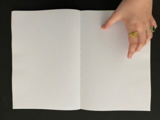 Eine Hand mit goldenen Ringen hält vor einem schwarzen Hintergrund ein leeres, weißes Notizbuch offen.