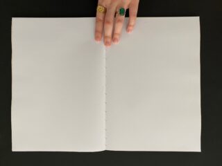 Eine Hand mit zwei Ringen hält auf einer dunklen Oberfläche ein leeres, spiralgebundenes Notizbuch offen.