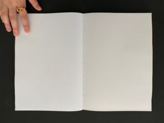 Eine Hand hält ein leeres, weißes, geheftetes Heft auf einer schwarzen Oberfläche offen.