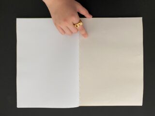 Eine Hand mit einem goldenen Ring zeigt auf die Mittelnaht eines offenen Buches mit leeren weißen und cremefarbenen Seiten auf einer schwarzen Oberfläche.