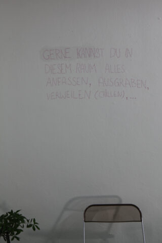 Eine weiße Wand mit deutschem Text in Rot, ein Metallstuhl und ein Teil einer grünen Pflanze in der unteren linken Ecke.