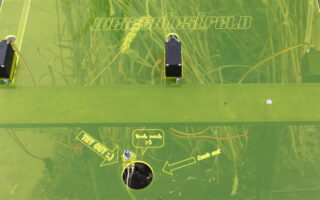 Eine grün getönte, transparente Platte mit elektronischen Komponenten und deutschen Texteinlagen, die über der Vegetation angebracht ist. Pfeile und Beschriftungen weisen auf Teile auf der Plattenoberfläche hin.