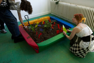 Zwei Personen kümmern sich um Pflanzen in einem kleinen, farbenfrohen Sandkastengarten im Innenbereich mit beschrifteten Markierungen auf einem grünen Boden in der Nähe eines Heizkörpers.