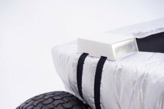 Eine Nahaufnahme eines metallischen, silberumwickelten Objekts, das mit zwei schwarzen Riemen neben einem großen Gummireifen befestigt ist, auf dem sich eine kleine weiße rechteckige Schachtel befindet.