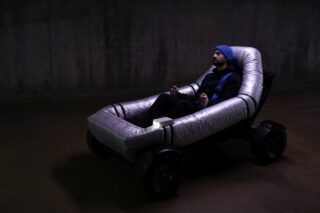 Eine Person mit einer blauen Mütze sitzt in einem metallischen, gepolsterten Stuhl auf Rädern in einem schwach beleuchteten Raum mit einer Betonwand.