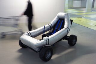 Ein vierrädriges Fahrzeug mit offenem Rahmen, aufblasbarem Sitz und Gurt ist in einem Gebäude geparkt; im Hintergrund ist unscharf eine Person mit einem Einkaufswagen zu sehen.