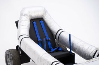 Ein schwarzer Sitz mit blauen Hosenträgergurten ist in einem mit silbernem Isoliermaterial überzogenen Rahmen montiert, der an einem kleinen Untergestell mit Rädern befestigt ist.