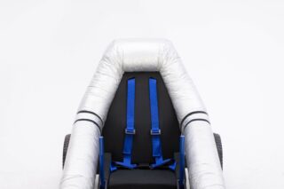 Eine Nahaufnahme eines minimalistischen Autositzes mit silbernem Rohrrahmen, schwarzem Kissen und blauen Hosenträgergurten vor einem schlichten weißen Hintergrund.