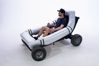 Ein Mann sitzt auf einem einfachen weißen Hintergrund in einem kleinen vierrädrigen Wagen mit aufblasbarem silbernen Rahmen, hält den Lenker in der Hand und ist angeschnallt.