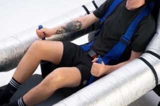 Eine Person in schwarzem Hemd und Shorts sitzt mit blauen Gurten in einem Sitz festgeschnallt und hält blaue Griffe neben großen silbernen isolierten Schläuchen.