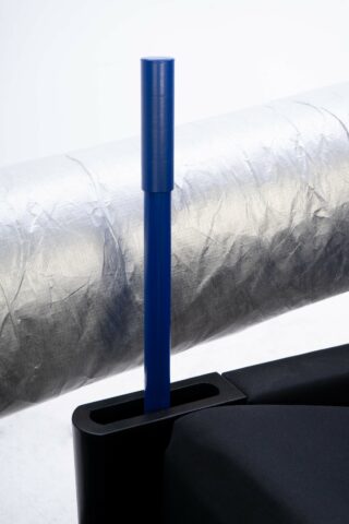 Ein blaues zylindrisches Objekt steht aufrecht auf einem rechteckigen schwarzen Sockel, im Hintergrund ist eine große silberne Metallrolle zu sehen.