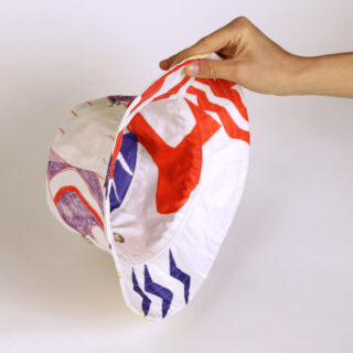 Eine Hand hält einen weißen Anglerhut mit abstrakten Mustern in Rot, Lila und Blau vor einem schlichten weißen Hintergrund.