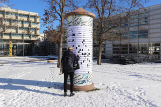 Eine Person mit einem Rucksack steht vor einer zylindrischen Außenausstellung, die mit kleinen weißen Tafeln bedeckt ist und von Schnee und modernen Gebäuden umgeben ist.