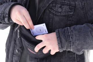 Eine Person in einer schwarzen Jeansjacke steckt eine Karte mit der Aufschrift „AT LEAST YOU ARE THERE“ in die Vordertasche einer schwarzen Gürteltasche.