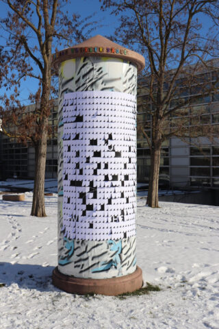 Eine zylindrische Litfaßsäule im Freien ist mit einem großen schwarz-weißen Kreuzworträtsel bedeckt, ringsherum sind Schnee und kahle Bäume zu sehen.