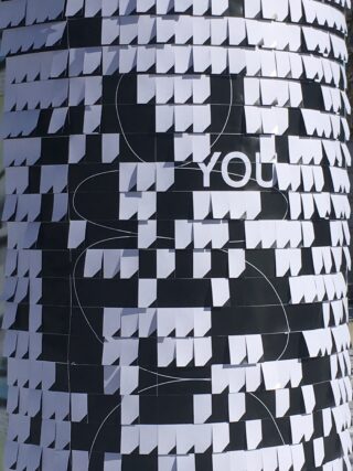 Eine zylindrische Oberfläche, die mit schwarzen und weißen Papierquadraten bedeckt ist, wobei auf einem Abschnitt das Wort „YOU“ in weißen Buchstaben aufgedruckt ist.
