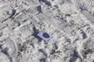 Eine Karte liegt auf schneebedecktem Boden mit sichtbaren Fußabdrücken und Grasflecken. Auf der Karte sind ein großer schwarzer Punkt und etwas gedruckter Text zu sehen.