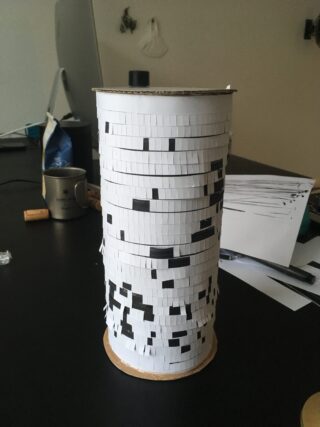 Ein zylindrisches Objekt, das mit weißem Papier mit schwarzen quadratischen Mustern bedeckt ist, steht auf einem dunklen Tisch, umgeben von Büromaterial und Papier.