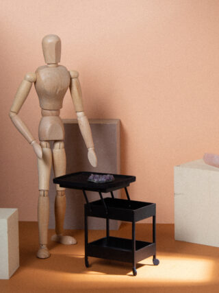 Eine hölzerne Künstlerpuppe steht neben einem kleinen schwarzen Rollwagen mit einem violetten Kristall darauf, umgeben von beigen geometrischen Blöcken.