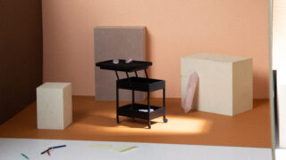 Zwischen drei rechteckigen Blöcken steht ein schwarzer Rollwagen mit zwei Regalen; auf dem Boden liegen verstreut Stifte und Papier in einer sanft beleuchteten, minimalistischen Umgebung.