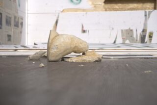 Eine zerbrochene Büste liegt in Einzelteilen auf dem Boden vor einer mit Paneelen und Zeitungspapier bedeckten Baustellenmauer.