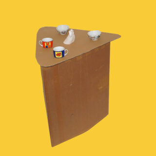 Eine dreieckige Pappstruktur mit vier Tassen und einer kleinen weißen Figur darauf, vor einem einfarbig gelben Hintergrund.