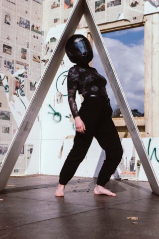 Eine Person mit schwarzem Helm und dunkler Kleidung steht barfuß unter einer dreieckigen Metallkonstruktion in einem Raum mit Zeitungspapier bedeckten Wänden und Graffiti.