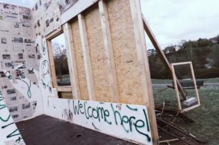 Im Freien steht eine teilweise errichtete Holzkonstruktion mit Zeitungspapier bedeckten Wänden und dem Graffiti „Willkommen hier!“, daneben liegen Trümmer und ein offener Fensterrahmen.