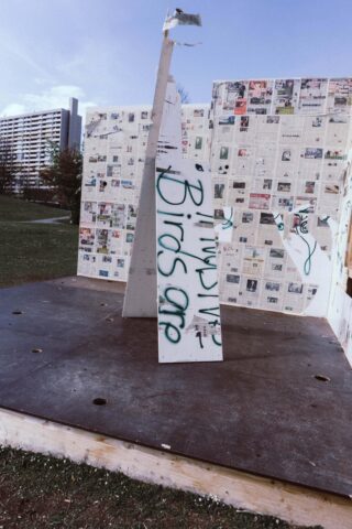 Eine Holzkonstruktion im Freien ist mit Zeitungsständen bedeckt. In der Mitte befindet sich eine Tafel mit dem grünen Graffiti „Birds are“. Im Hintergrund sind Wohnhäuser und Gras zu sehen.
