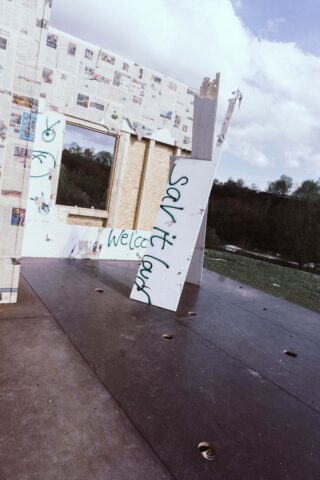 Teilweise errichtete, mit Zeitungspapier bespannte Wand mit einer seitlich stehenden Tür, besprüht mit den Aufschriften „Sanitarium“ und „Willkommen“. Im Hintergrund ein grasbewachsener Hügel und ein bewölkter Himmel.