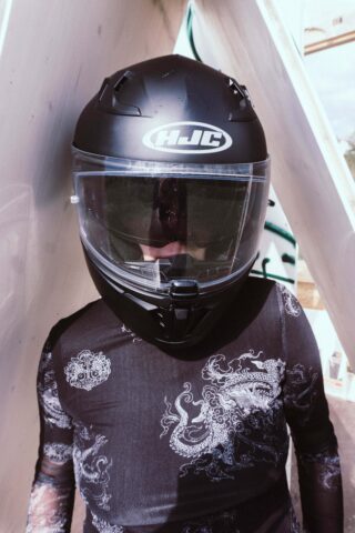 Eine Person mit einem schwarzen HJC-Motorradhelm und einem schwarzen Hemd mit einem weißen Drachenmuster steht vor einem weißen Hintergrund mit Graffiti.