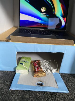 Ein Laptop steht auf einem hellblauen Karton mit einem ausgeschnittenen Fach, in dem sich Taschentücher, ein Twix-Riegel und ein Paar weiße Kopfhörer befinden.