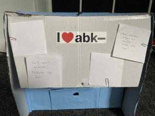 Ein Pappkarton mit einem Schild mit der Aufschrift „I ♥ abk–“ und drei handgeschriebenen Notizen, die mit Büroklammern an der Vorderseite befestigt sind. Der Karton steht auf einem Teppichboden.