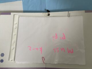 Ein umgedrehtes Stück Papier mit rosa handgeschriebenem Text, auf dem teilweise „Miss“ und „Pipi“ zu lesen sind, ist mit Büroklammern und Klebeband an einem Ordner befestigt.