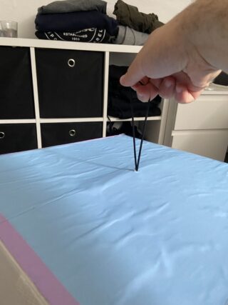 Eine Hand zieht ein Gummiband durch ein Loch in einer blauen Abdeckung, die auf einer Kiste in einem Raum mit Regalen und gefalteter Kleidung im Hintergrund liegt.