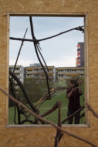 Eine Person steht hinter einem Holzrahmen mit sich kreuzenden Ästen, im Hintergrund sind Wohnhäuser und Grünflächen zu sehen.
