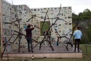 Drei Personen interagieren mit einer großen Holzstabkonstruktion vor mit Zeitungsblättern bedeckten Wänden im Freien auf einer Rasenfläche. In der Nähe steht eine Flasche auf dem Boden.