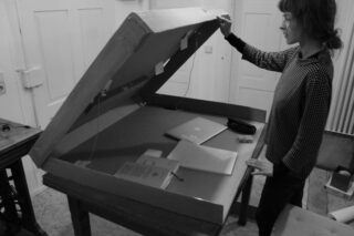 Eine Person steht an einem Tisch und hebt den großen Deckel eines Kartons an. Darin befinden sich ein geschlossener Laptop, ein Notizbuch, Papiere und ein Buch.