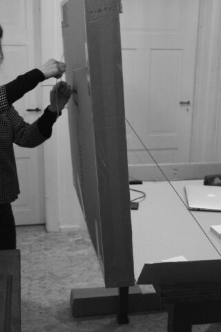 Eine Person hält und zieht an einer Schnur, die an einer großen Pappplatte befestigt ist, die aufrecht auf einem Tisch in einem Raum mit geschlossenen Türen steht.