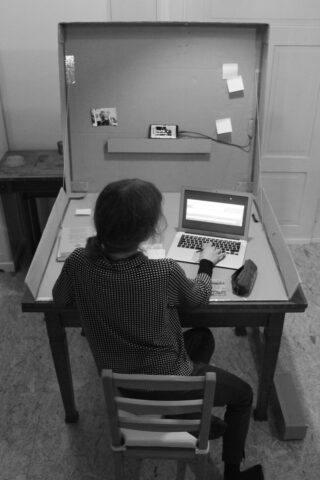 Eine Person sitzt an einem Schreibtisch und arbeitet an einem Laptop. Auf dem Schreibtisch in einem minimalistischen Arbeitsbereich liegen Papiere, Haftnotizen und ein Federmäppchen.