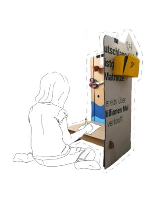 Ein Kind sitzt auf dem Boden und zeichnet auf Papier vor einer Pappkonstruktion mit deutschem Text und kleinen Regalen mit Kunstbedarf.