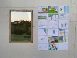 Ein Fenster mit Holzrahmen an einer Wand neben einem Raster aus sechzehn handgezeichneten und gemalten Bildern, die Landschaften, Gebäude und abstrakte Szenen darstellen.