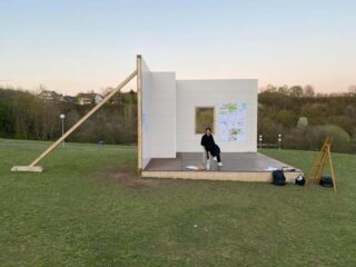 Eine Person sitzt auf einem Stuhl in einem halbfertigen Raum mit zwei Wänden und einem Fenster auf einer Wiese. An einer Wand sind Architekturzeichnungen befestigt.
