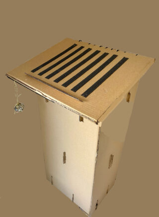 Eine Pappkartonfalle mit Schlitzdeckel und einem darin hängenden Köder vor einem schlichten Hintergrund.