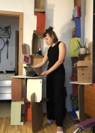 Eine Frau steht an einem provisorischen Stehpult aus Pappe und arbeitet an einem Laptop in einem mit Pappmöbeln und Pflanzen dekorierten Raum.