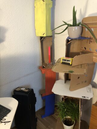 In einer Ecke stehen verschiedene Pappkonstruktionen, die ein gelbes Maßband, eine rote Kerze, Topfpflanzen und einige bemalte Tafeln enthalten.