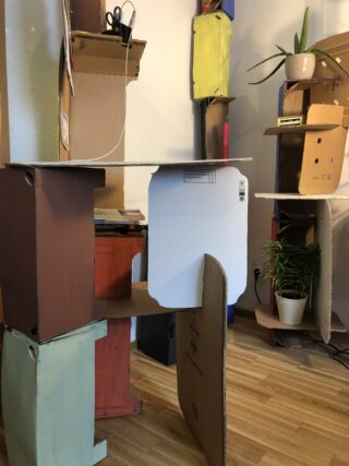 In einem Raum sind Möbel und Regale aus Pappe aufgebaut, in denen auf einem Holzboden verschiedene Gegenstände wie Pflanzen, Scheren und Büromaterial aufbewahrt werden.