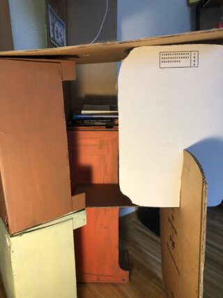 Auf einem Holzboden ist eine Konstruktion aus verschiedenen bemalten Kartons und Blättern gestapelt, an einer Platte ist eine kleine Sehtesttafel befestigt.
